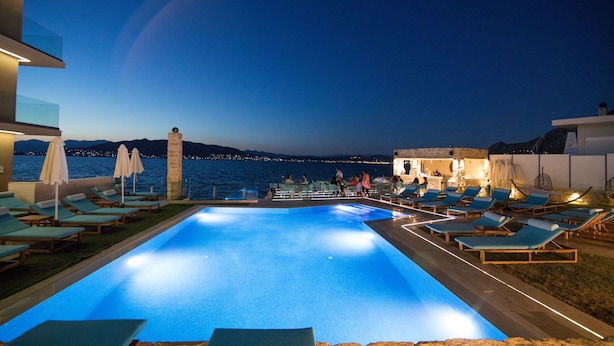Une piscine à débordement avec des chaises longues et des parasols, offrant une vue sur une étendue d’eau et les lumières de la ville au loin.