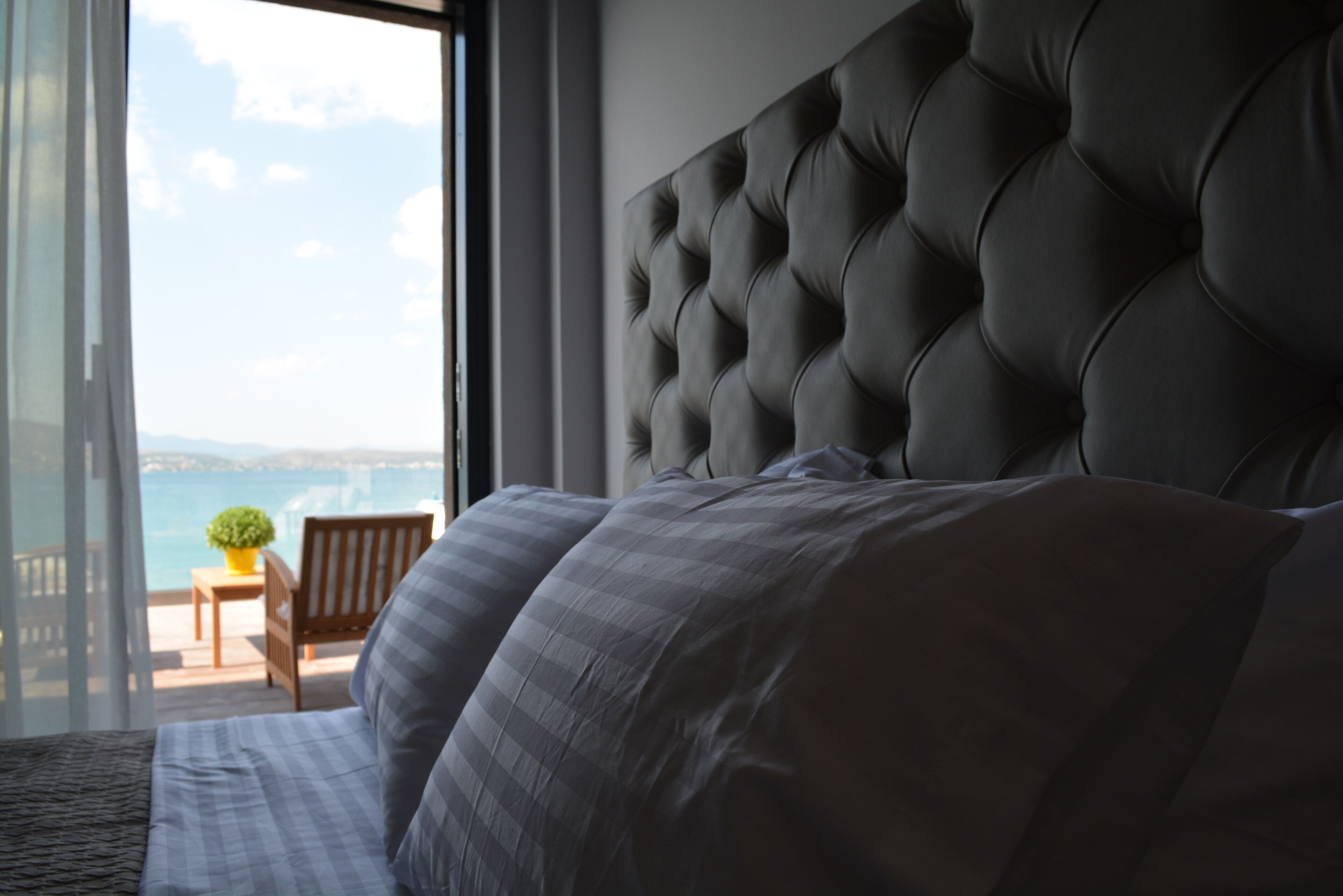 A bedroom with a large bed, a tufted headboard, a view of the sea, and a balcony with a chair and a plant.