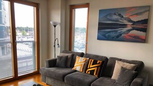 A modern living room with a grey sofa, a black coffee table, and a cactus decoration. There are large windows with blinds, a floor lamp, and a painting on the wall. - The Wharf Medway (Chatham)