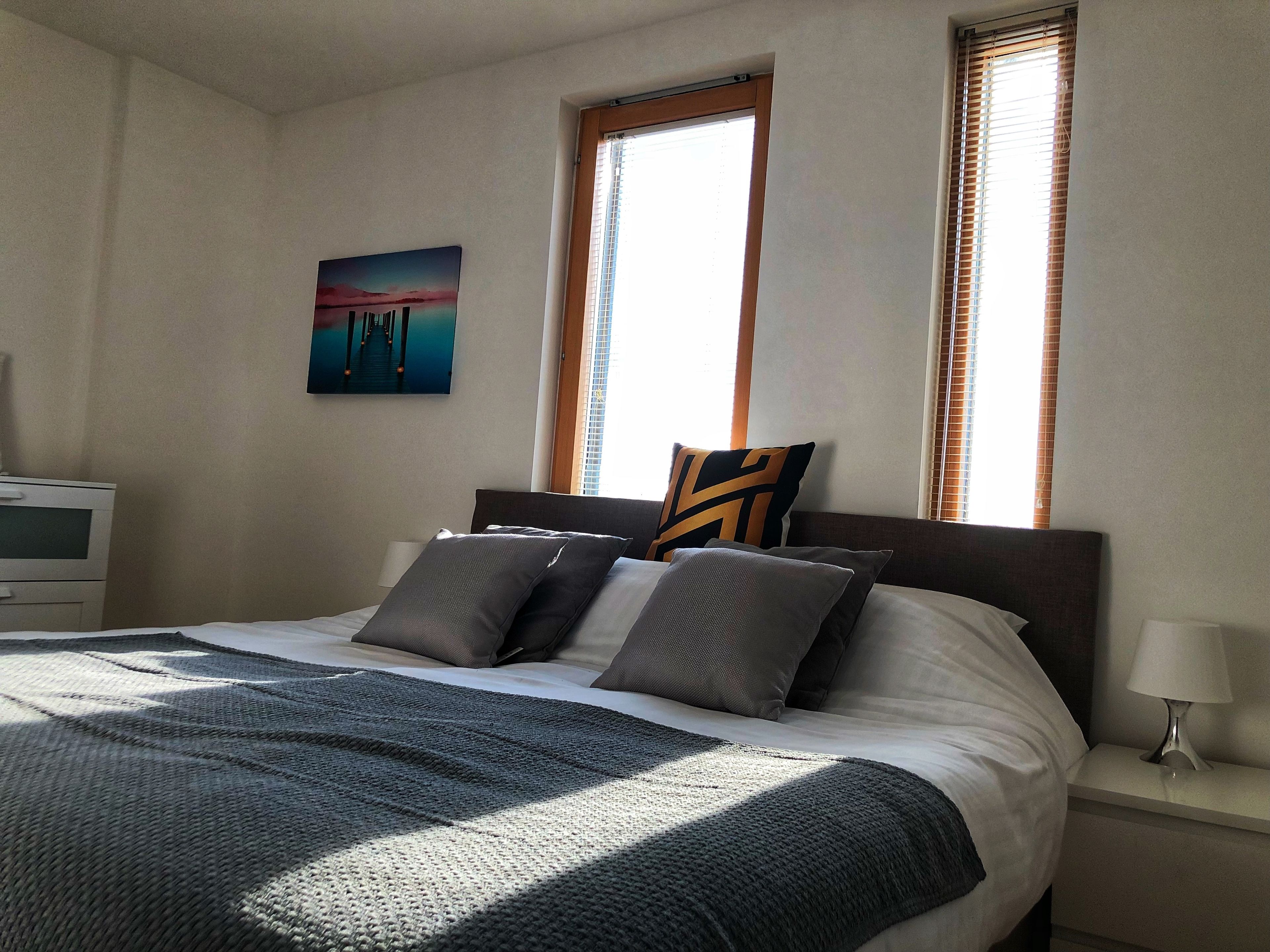 A neatly made bed with a gray blanket and pillows, a bedside lamp, and a painting on the wall.