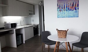 A modern kitchen with white cabinets, a microwave, and a sink. There is a white table with two chairs and a woven basket on it. - EcoApart Antofagasta (Antofagasta)