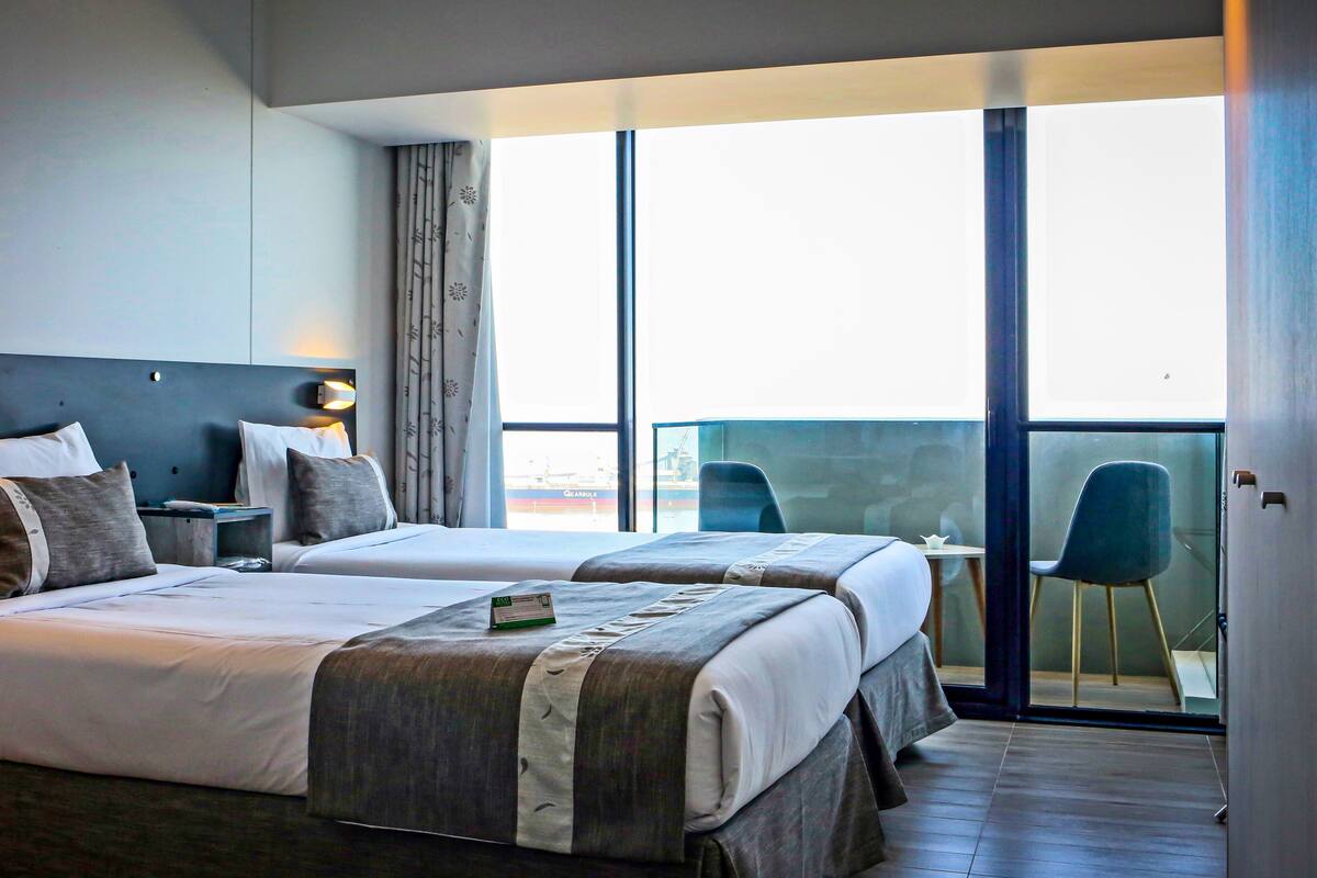 A hotel room with two beds, a small table, a chair, and a balcony with a view of the sea.