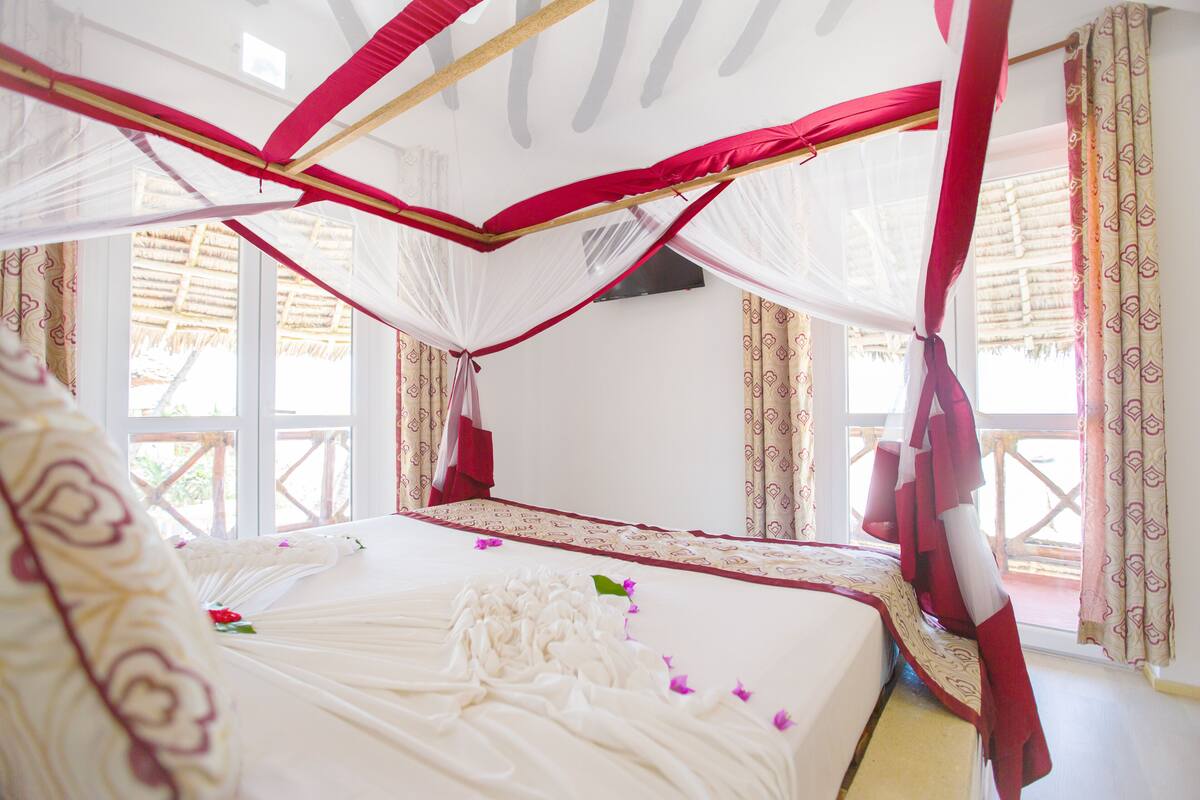 Un lit à baldaquin, une fenêtre donnant sur une structure à toit de chaume et un rideau à motifs.