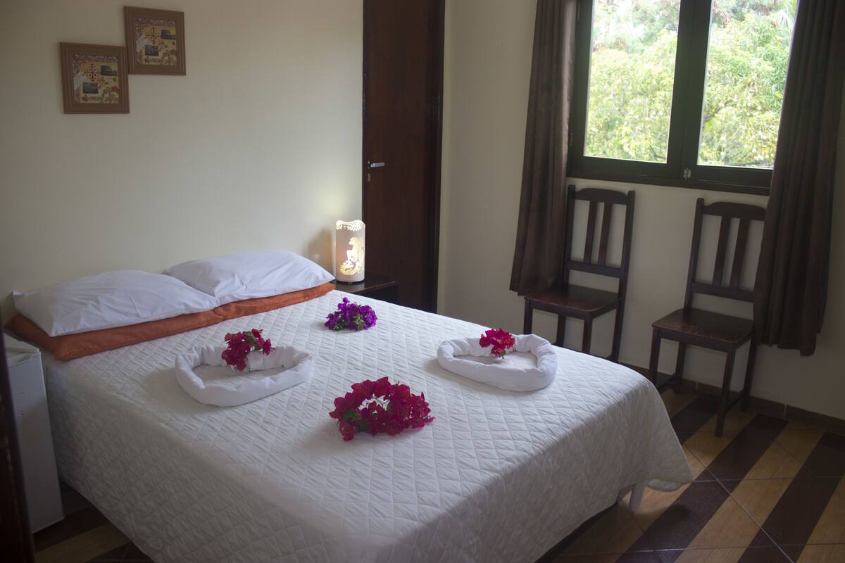 A neatly made bed with white linens and decorative flower arrangements, a wooden nightstand with a lamp, and two wooden chairs in a room with a window and curtains.
