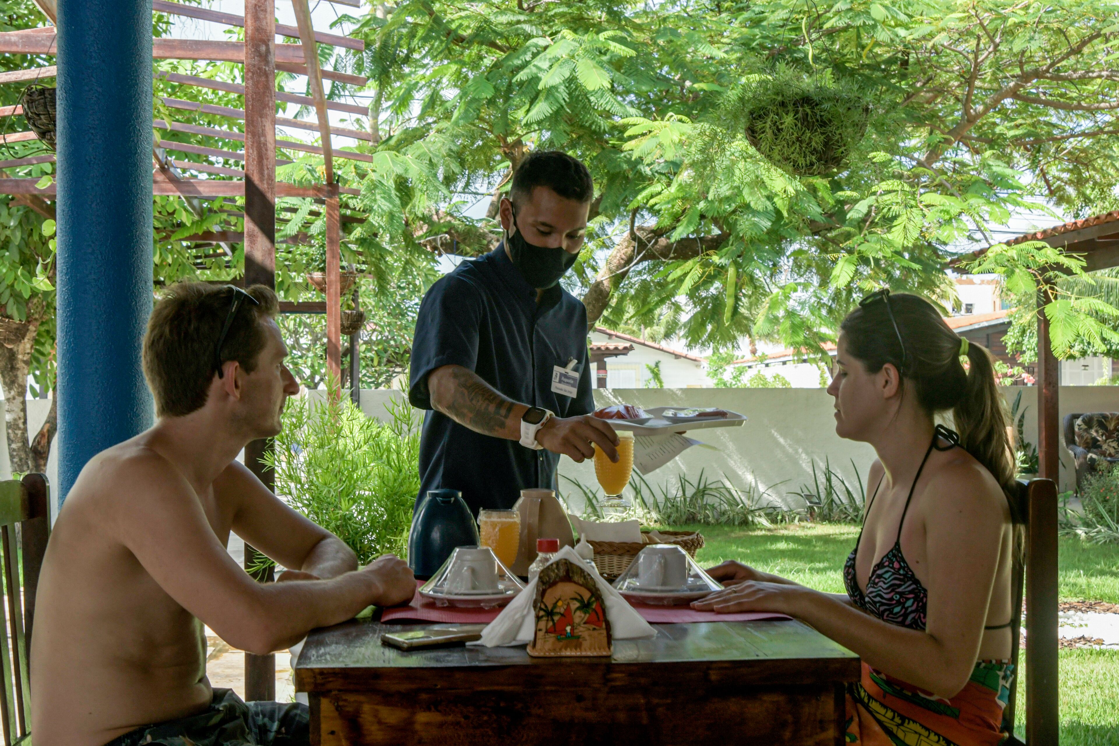 A dining area with a table set for two, a waiter serving a drink, and a tropical outdoor setting with trees and a wooden pergola.