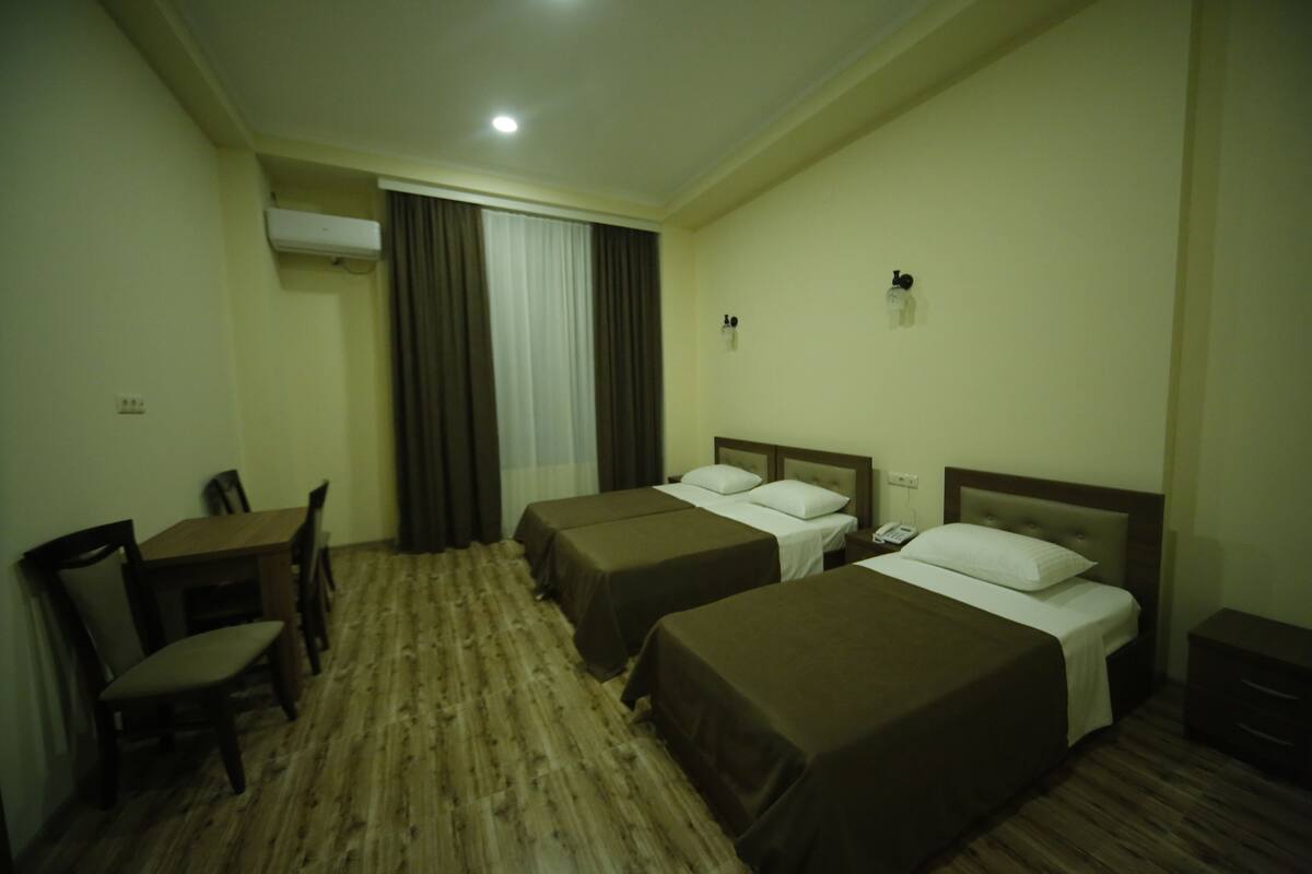 Ein Hotelzimmer mit zwei Einzelbetten, einem Schreibtisch mit Stuhl, einem kleinen Tisch und einem Fenster mit Vorhängen.