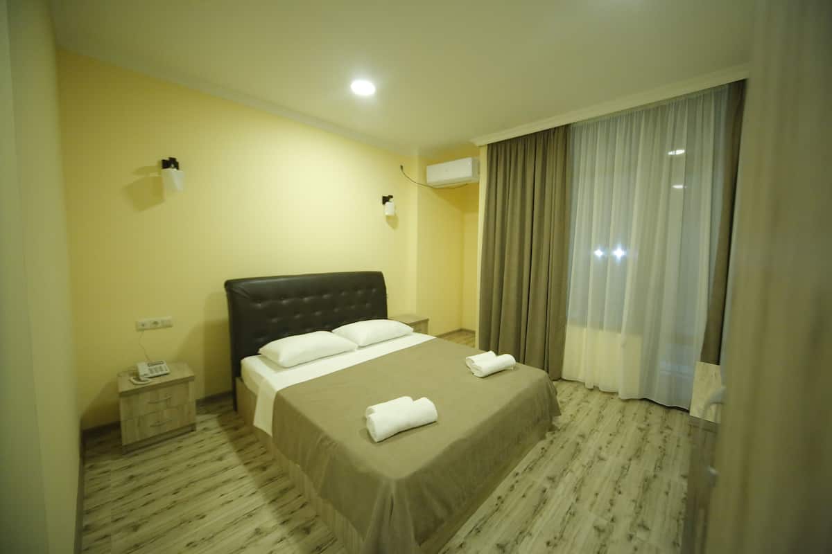 Ein Hotelzimmer mit einem großen Bett, einem Nachttisch mit Telefon, einer Klimaanlage und einem Fenster mit Vorhängen.