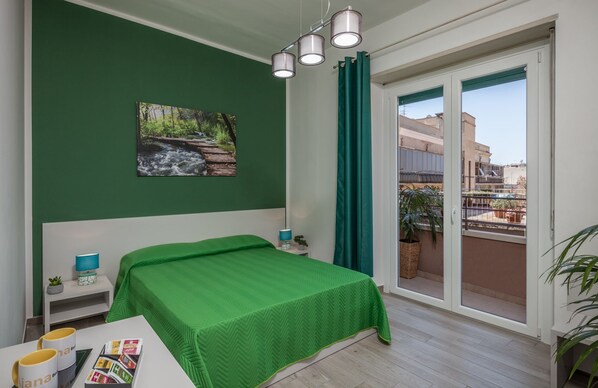 Un dormitorio con colcha verde, una mesita de noche con lámpara, una ventana con vista a edificios y un cuadro de una cascada.