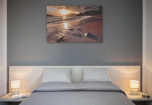 A bedroom with a bed, two pillows, a nightstand with a lamp, and a framed picture of a beach scene. - Ajana Rooms by Accomodo (Cagliari)