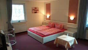 Een slaapkamer met een bed, nachtkastjes, een klein tafeltje, een radiator en een raam met gordijnen.