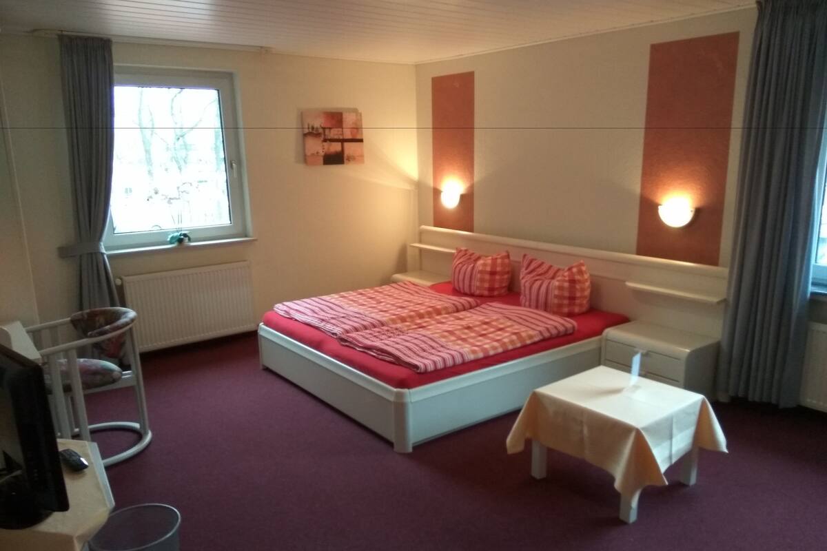 A bedroom with a bed, bedside tables, a small table, a radiator, and a window with curtains.