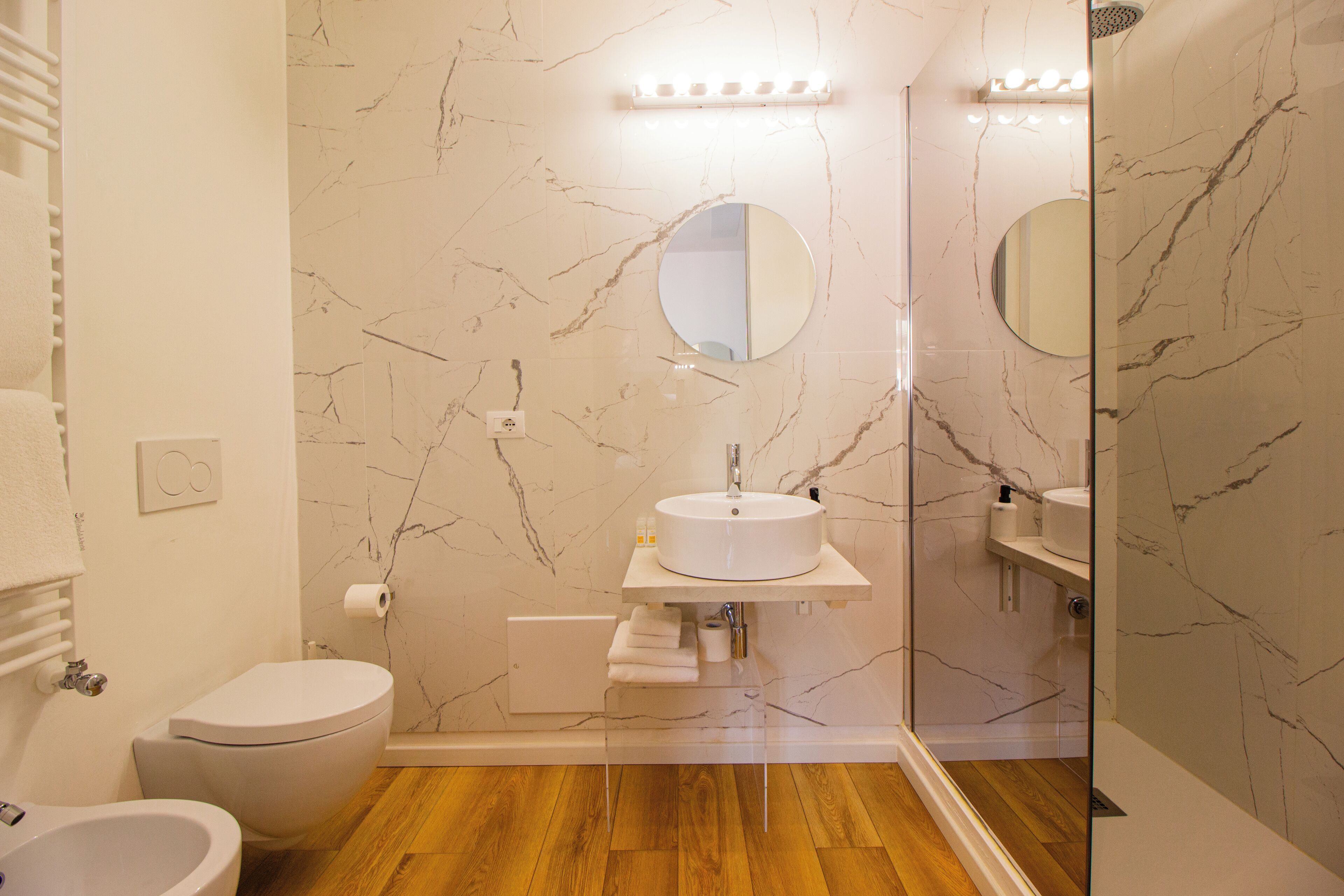 A modern bathroom with a marble backsplash, a round mirror, a white sink, and a toilet.