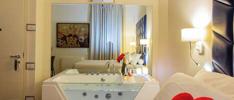 A hotel room with a large bed, a glass-enclosed bathtub, a painting on the wall, and a mirror reflecting the room.