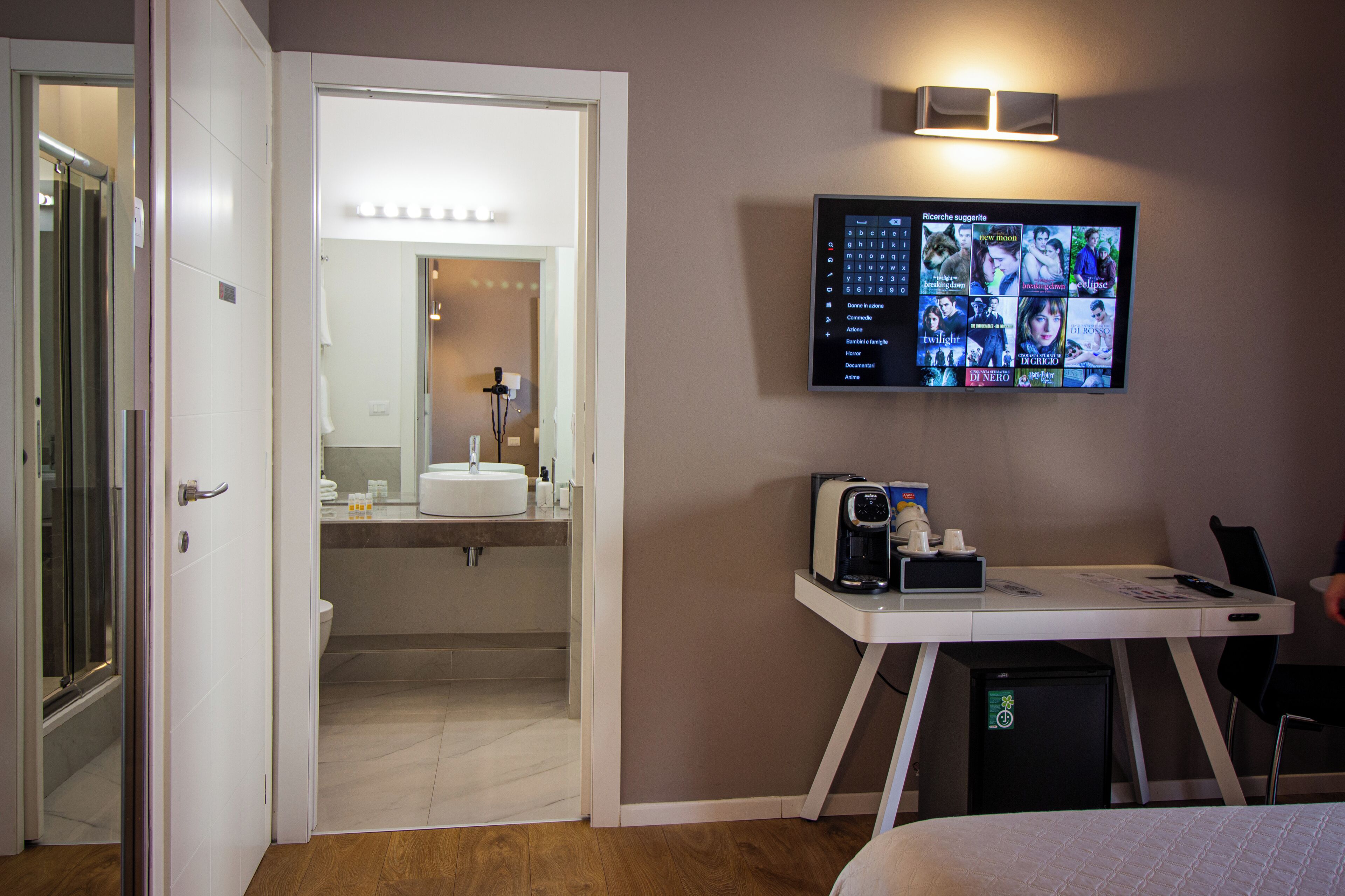 Una camera d'albergo con un letto, una scrivania con computer e macchina del caffè, una TV a parete che mostra una selezione di film, e un bagno visibile attraverso una porta aperta.