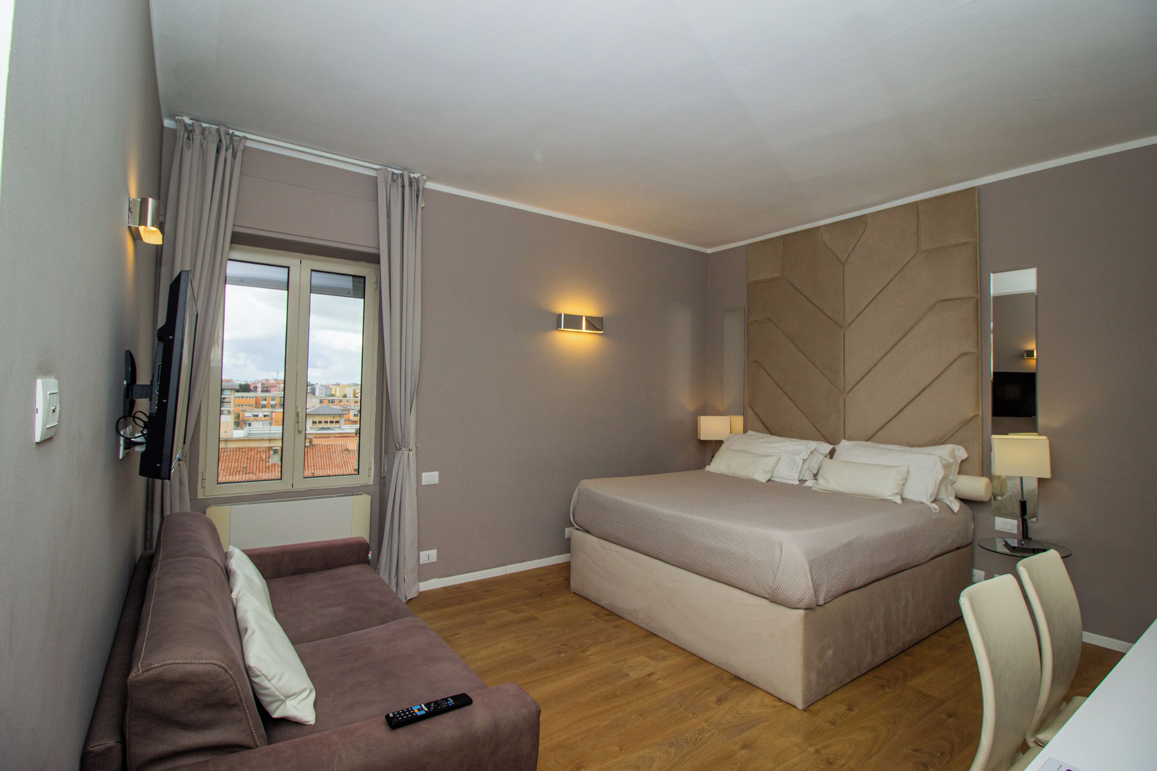 Habitación de hotel con cama, sofá, televisión, ventana con cortinas y vistas a edificios.