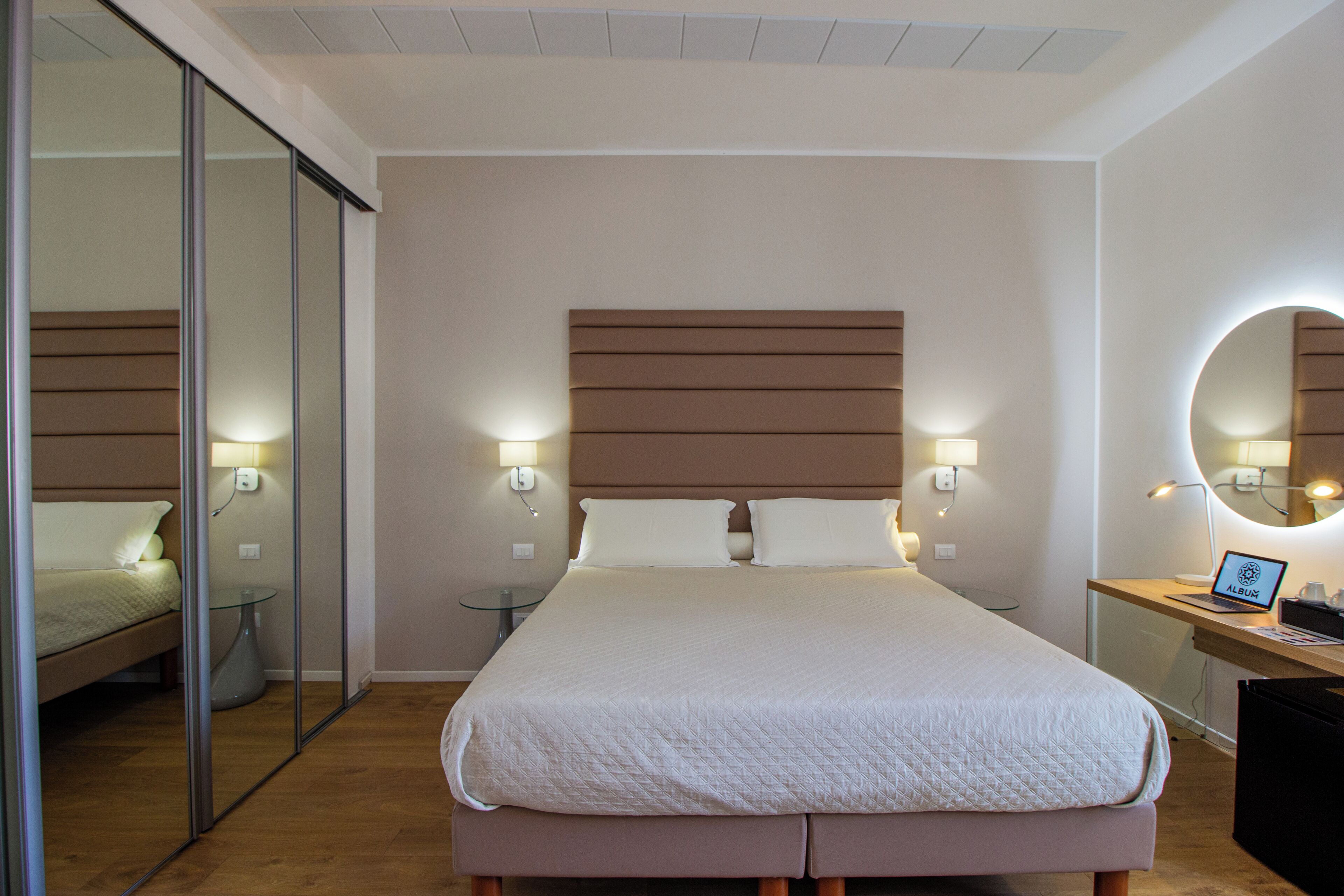 Una habitación de hotel moderna con una cama grande, lámparas de noche, un espejo redondo y un ordenador portátil sobre una mesita de noche de madera.