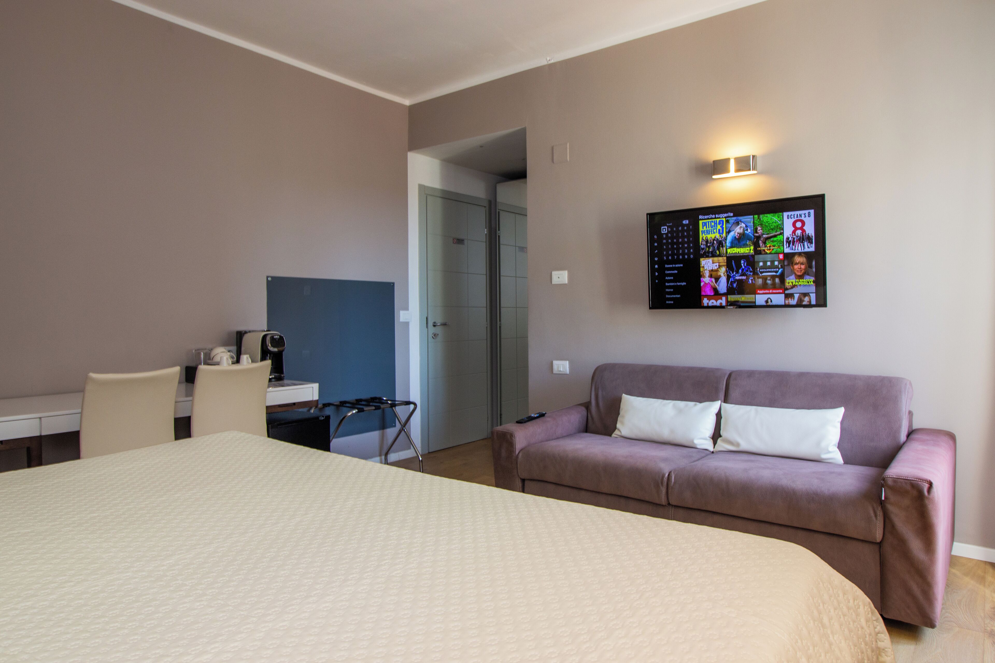 A hotel room with a large bed, a sofa, a desk with a chair, and a TV mounted on the wall displaying various channels.