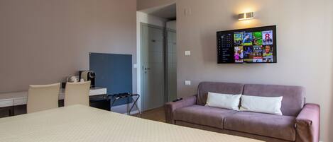A hotel room with a large bed, a sofa, a desk with a chair, and a TV mounted on the wall displaying various channels.