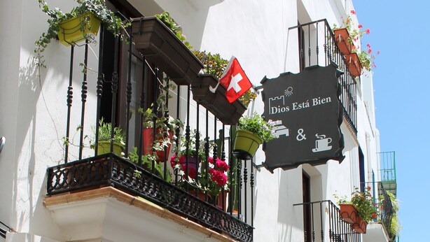 Un café con mesas al aire libre, un letrero que dice "Dios Está Bien" y plantas en macetas en el balcón.