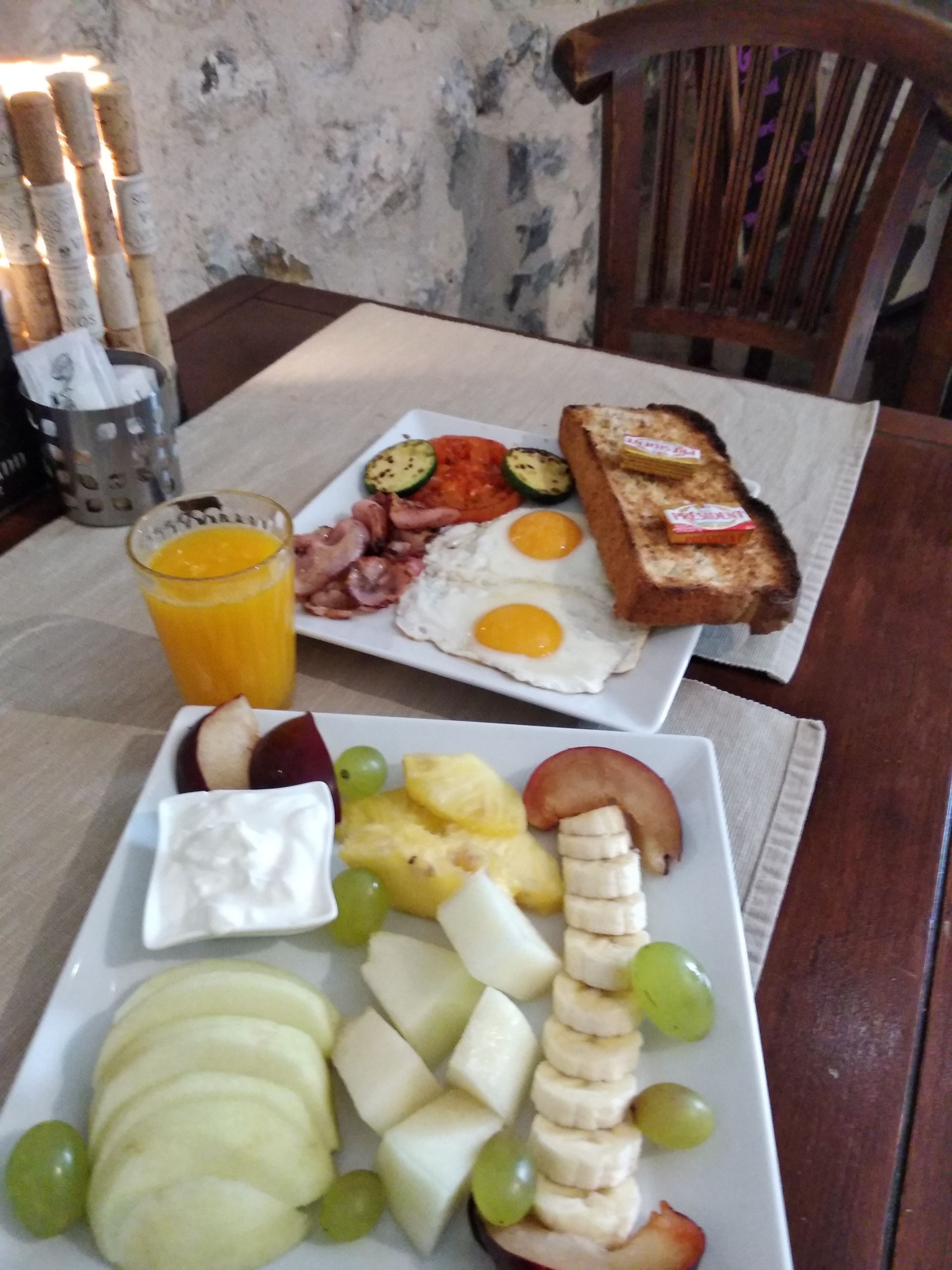 A breakfast plate with eggs, bacon, toast, and fruit, and a glass of orange juice on a table.