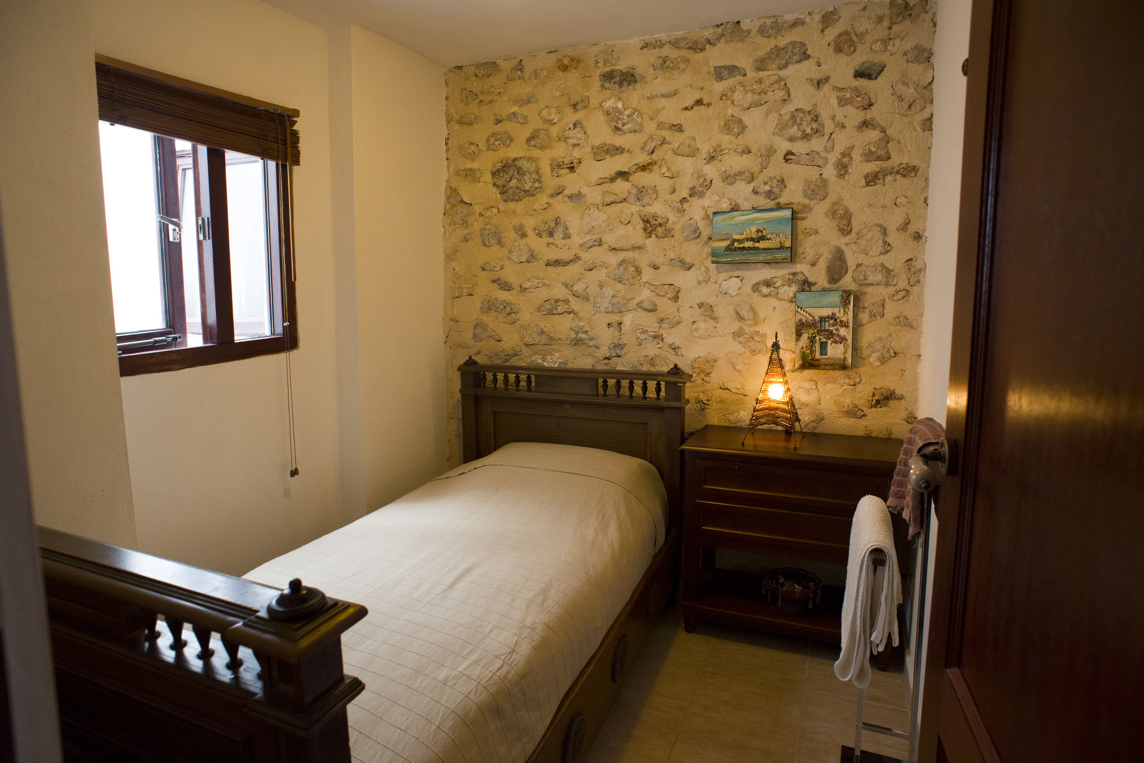 A bedroom with a single bed, a nightstand, a lamp, and a stone wall.