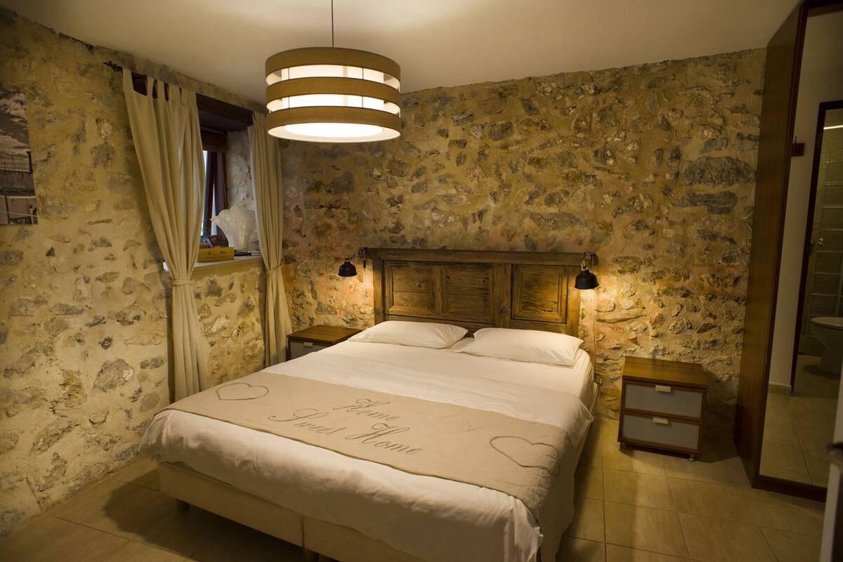 Quarto com parede de pedra, cabeceira de madeira, cama com colcha em formato de coração e mesa de cabeceira com abajur.