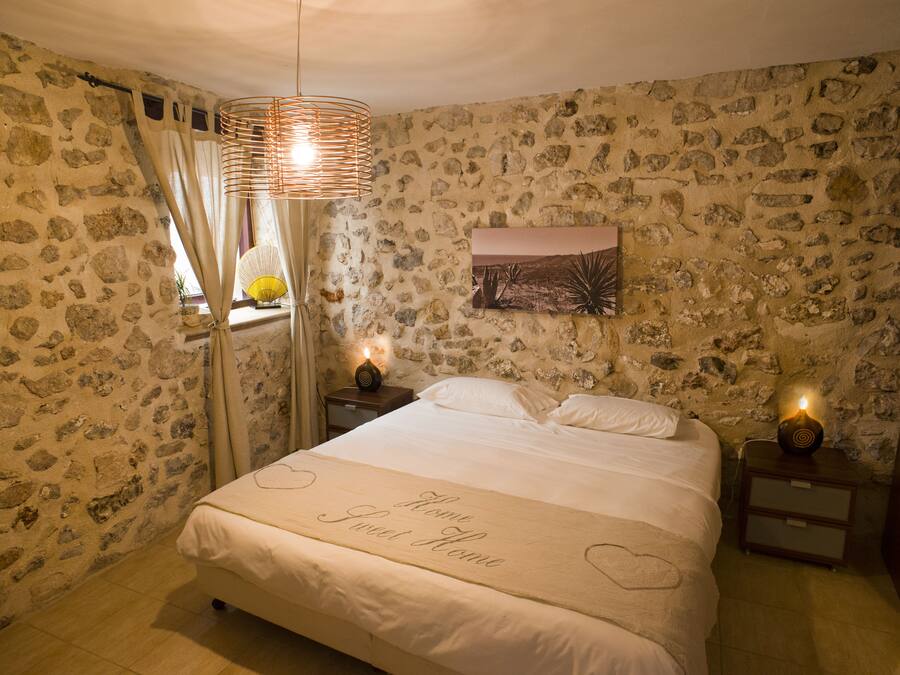 A bedroom with a stone wall, a bed with a heart-shaped bedspread, and a nightstand with a lamp.