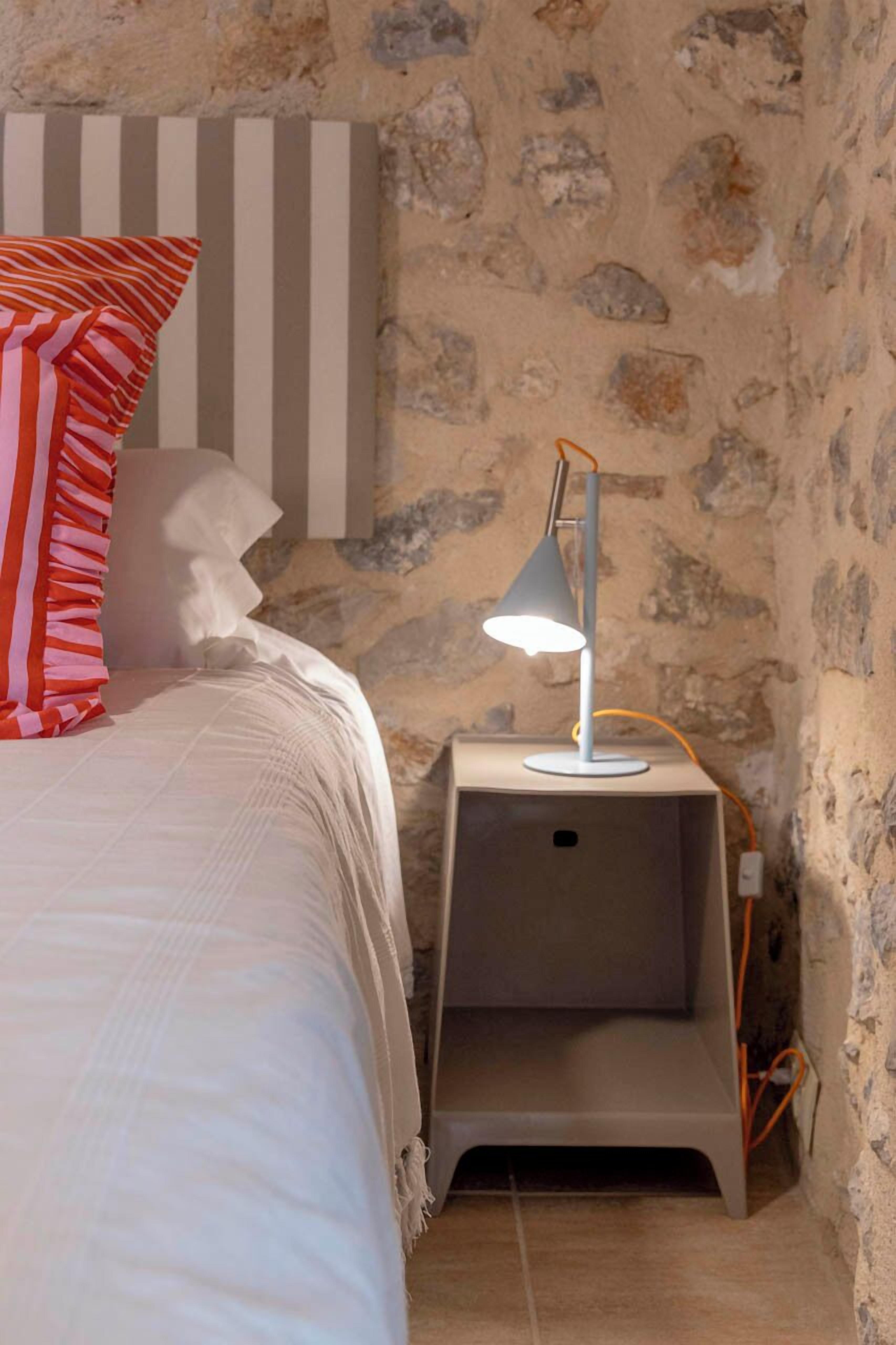 Un dormitorio con una cama, un cojín a rayas, una lámpara y una pared de piedra.