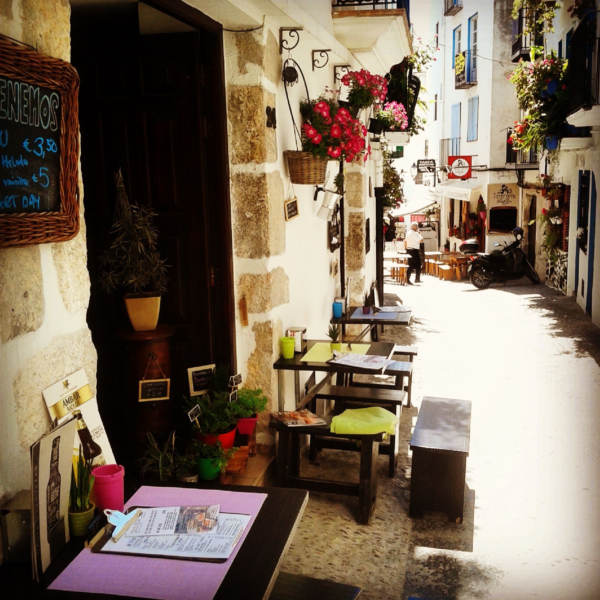A narrow street with outdoor seating, potted plants, and a signboard.