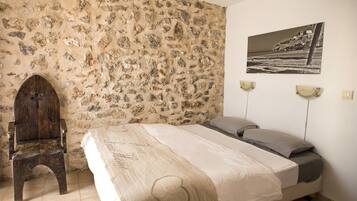 A bedroom with a stone wall, a wooden chair, a bed with a beige bedspread, and a black and white photograph on the wall.
