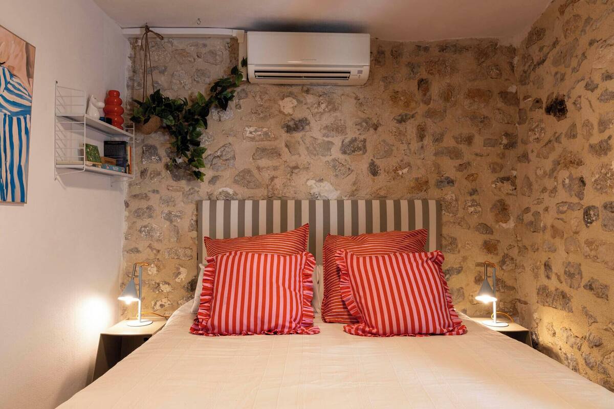 Quarto com parede de pedra, cama com almofadas listradas, ar condicionado e abajur.
