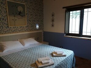 Een net opgemaakt bed met een beddekleed met patroon en twee gevouwen handdoeken, een wandklok en een raam met uitzicht op groen.