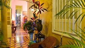 A hallway with checkered flooring, potted plants, wicker furniture, and a window with bars. - Hostal D' Cordero (Santa Clara)