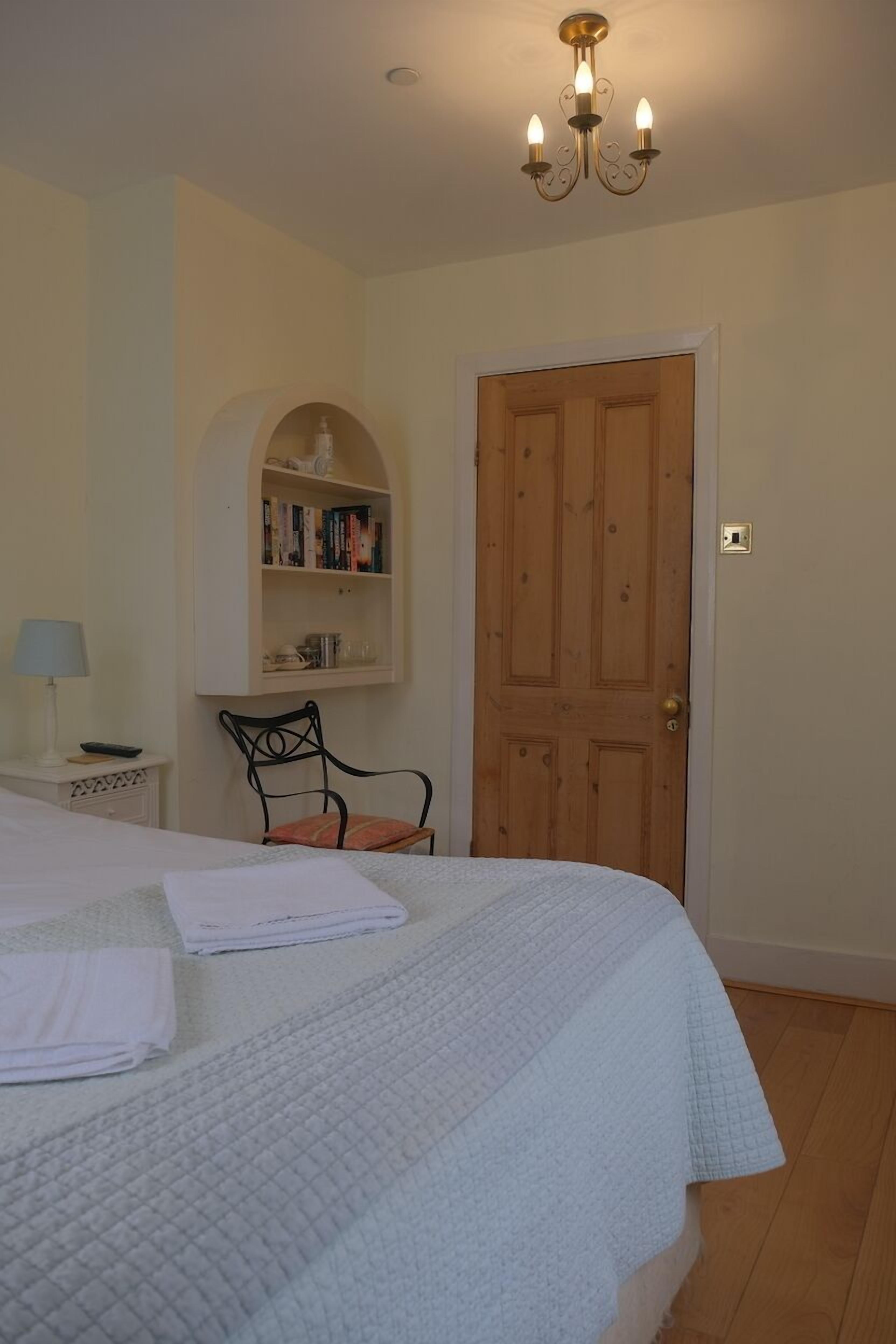 Een slaapkamer met een bed, nachtkastje, stoel, boekenkast en een houten deur.