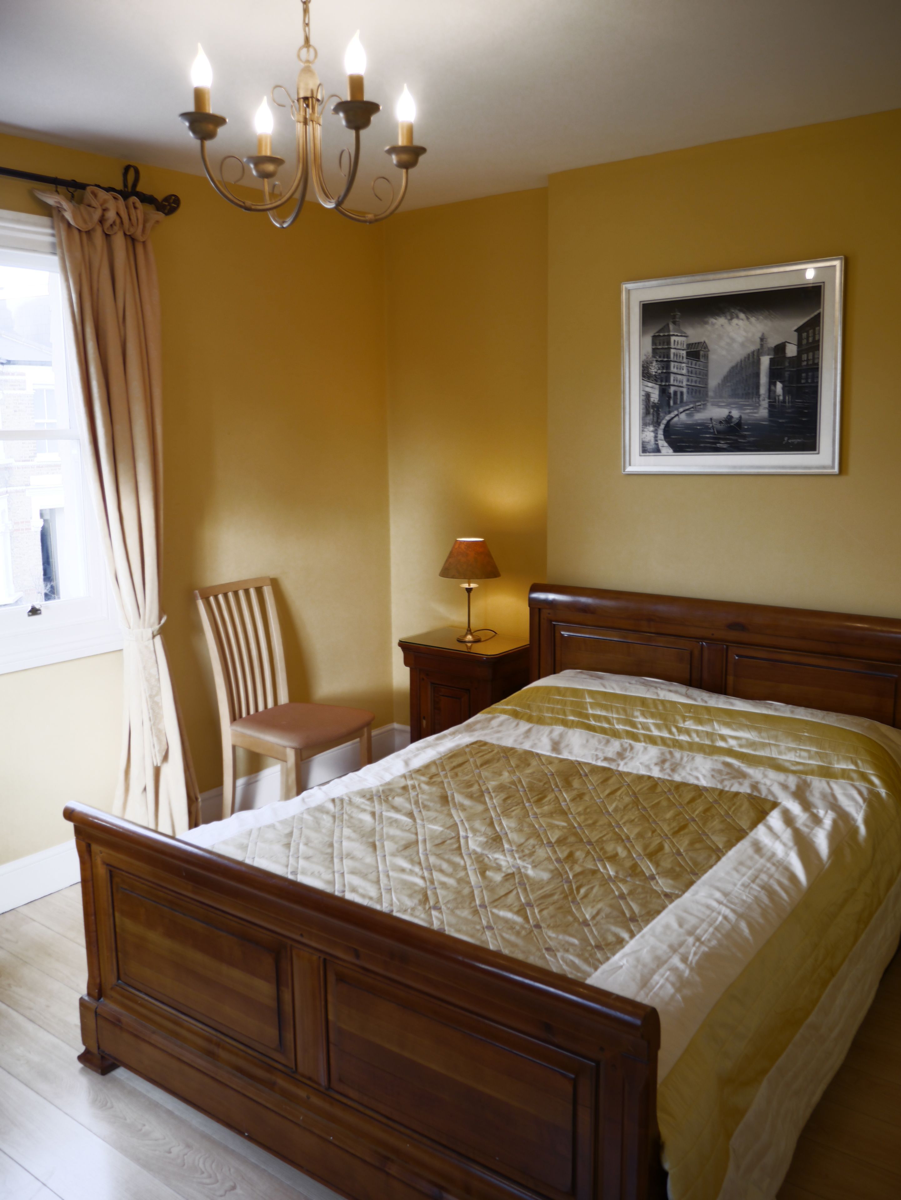 A bedroom with a wooden bed, a chair, a nightstand with a lamp, a wall-mounted picture, and a window with curtains.