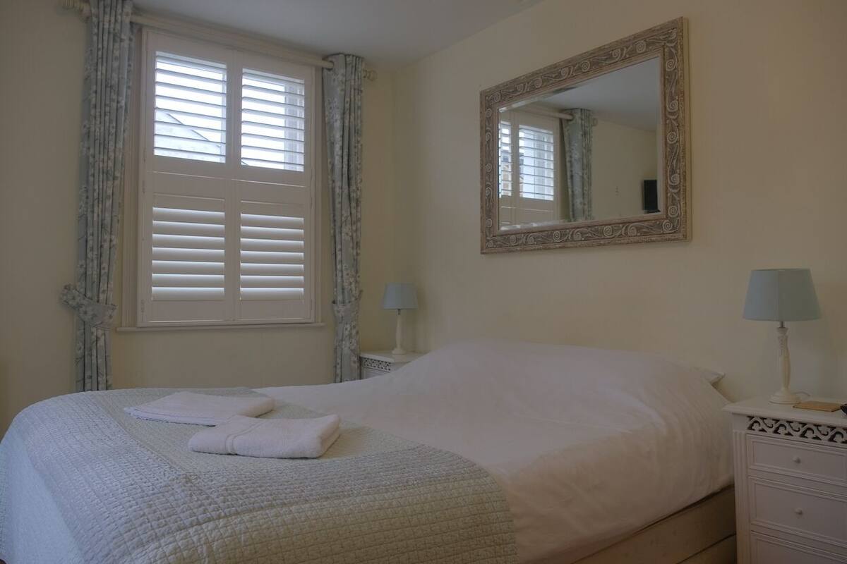 A neatly made bed with white linens and a folded towel, a bedside table with a lamp, a mirror on the wall, and a window with blinds.