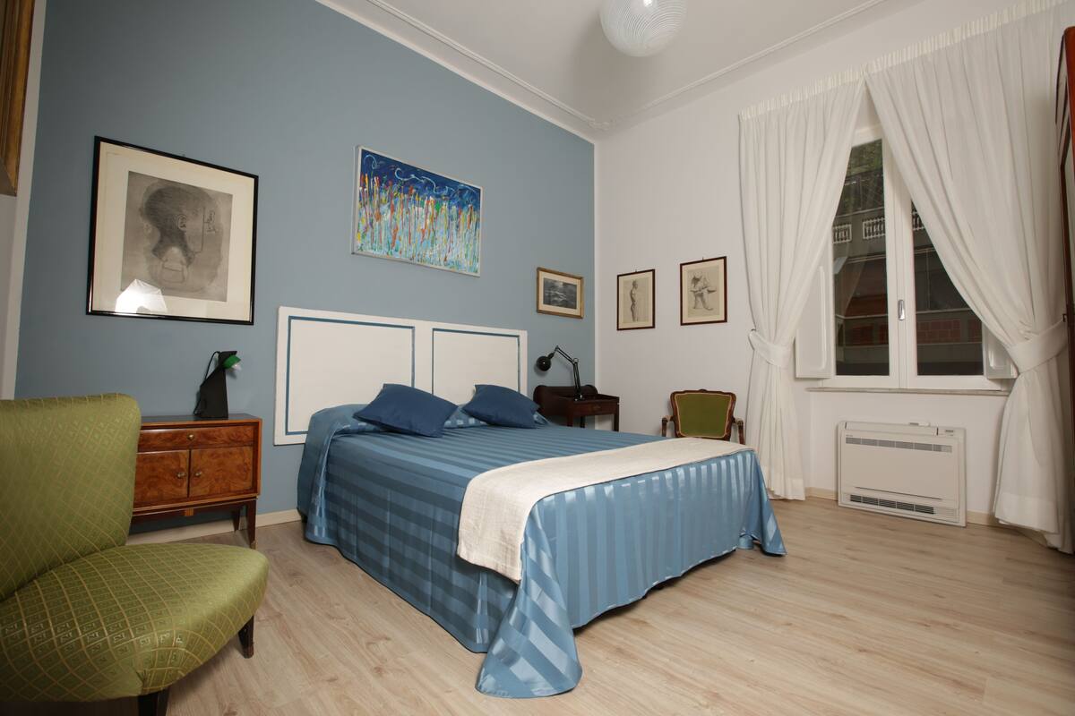 Un dormitorio con cama, una silla, una mesita de noche y una ventana con cortinas.
