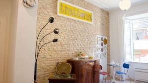 Una habitación con pared de ladrillo, un reloj, un cartel enmarcado, una mesa con sillas y una ventana con persianas.