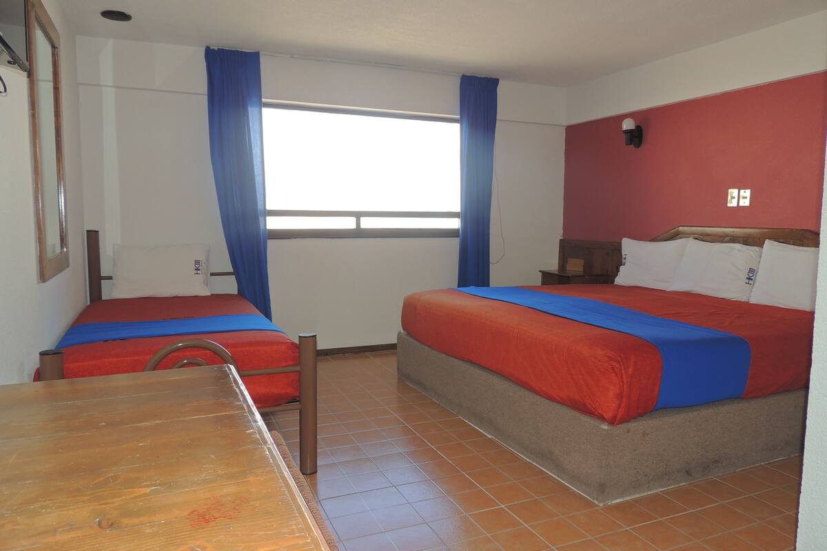 A room with two beds, a wooden table, a window with blue curtains, and a red wall.