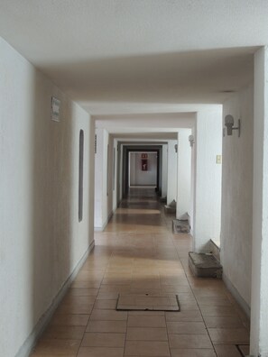 Um corredor comprido e estreito com paredes brancas e chão de azulejos. Há várias portas de acesso a quartos, e um extintor de incêndio fixado na parede.