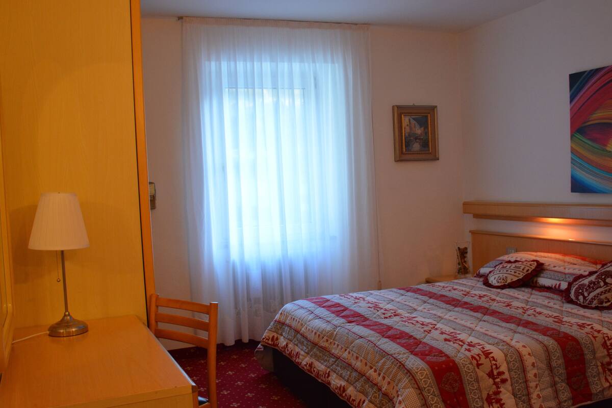 Hotellihuone, jossa on sänky, työpöytä, tuoli, lamppu, ikkuna verhoilla ja taulu seinällä.