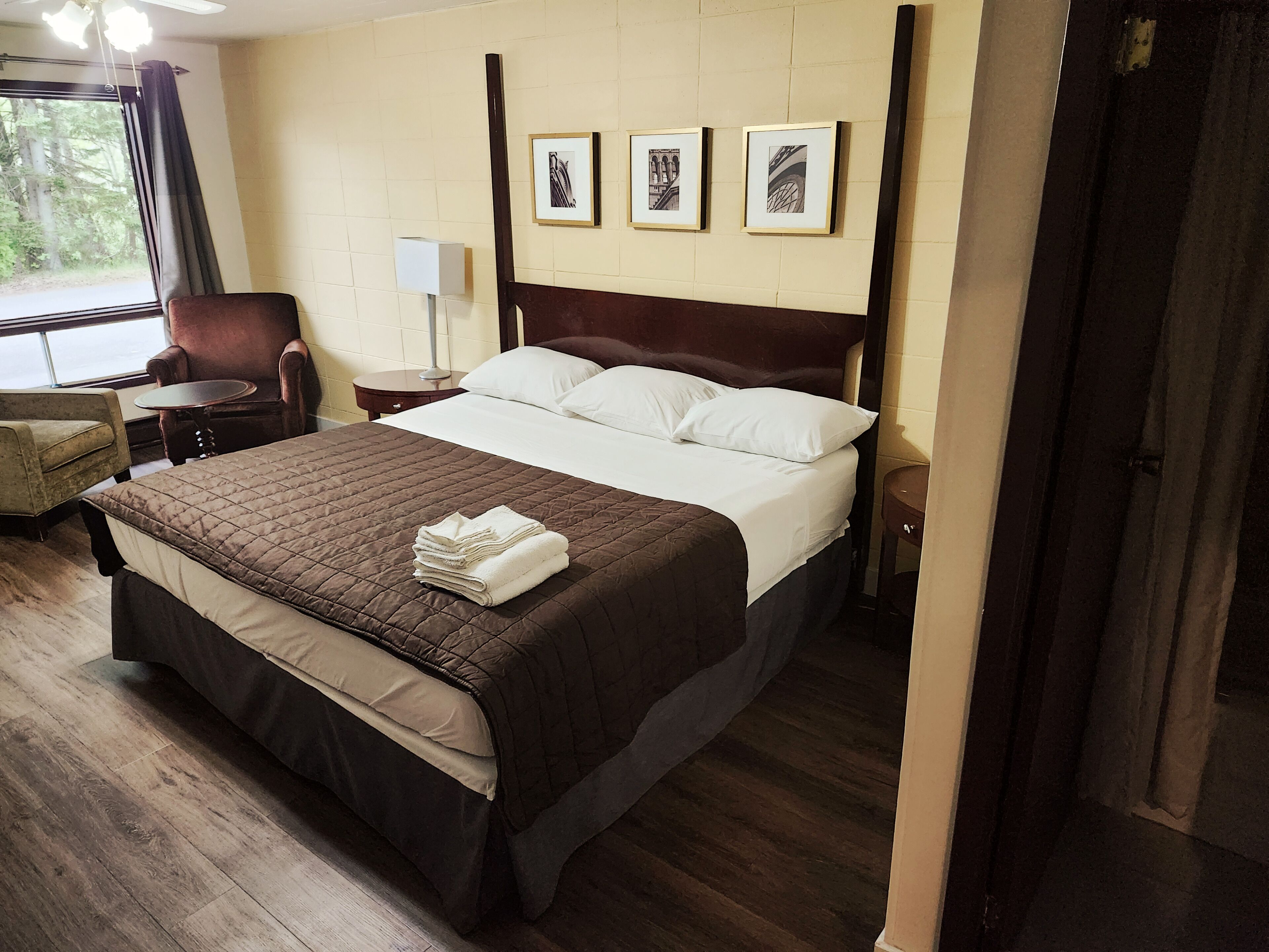 A neatly made bed with a brown quilt, a chair, a bedside table, and a lamp. There are framed pictures on the wall.
