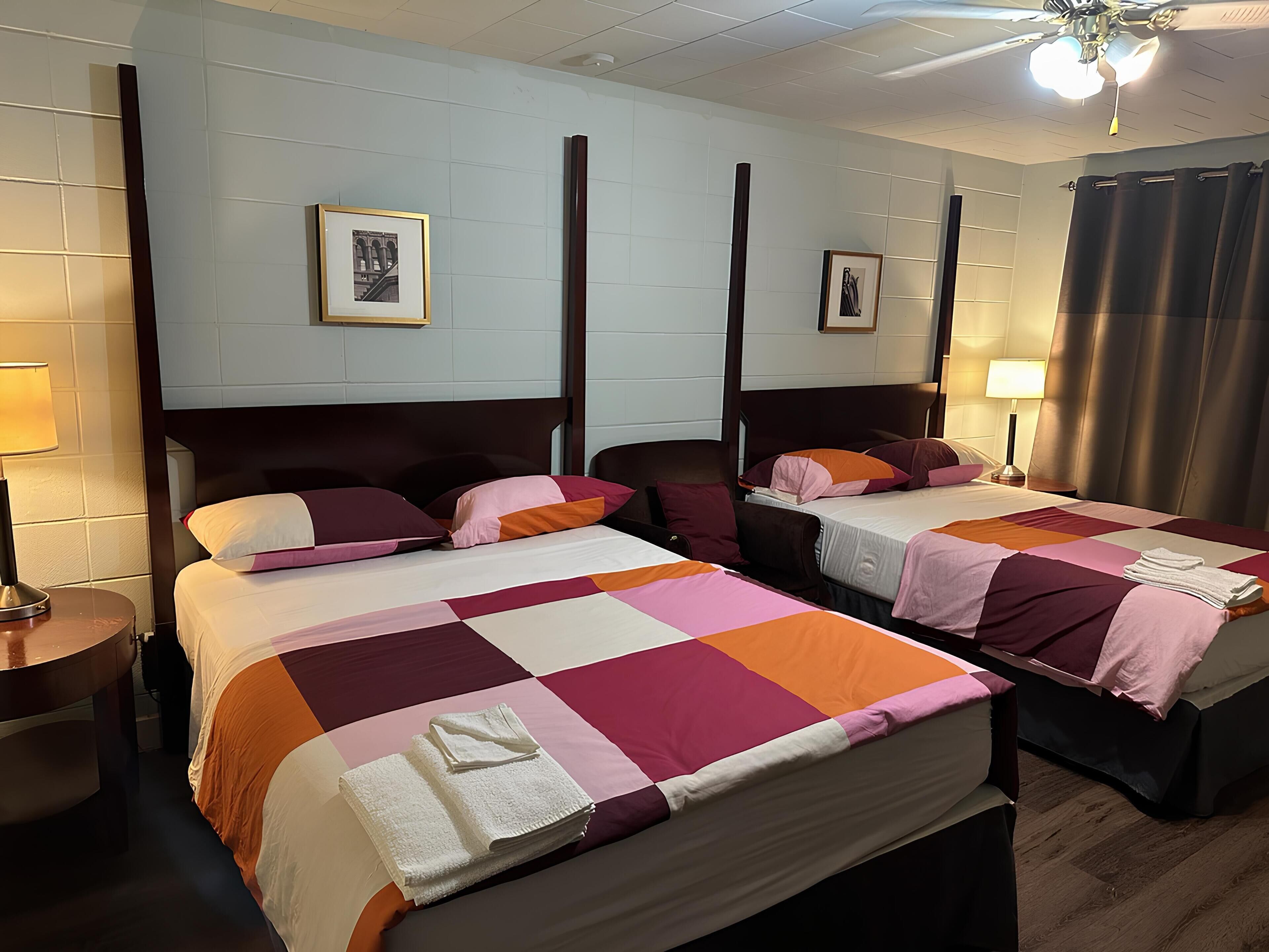 A hotel room with two beds, each with a wooden headboard, a large window with curtains, and a ceiling fan.