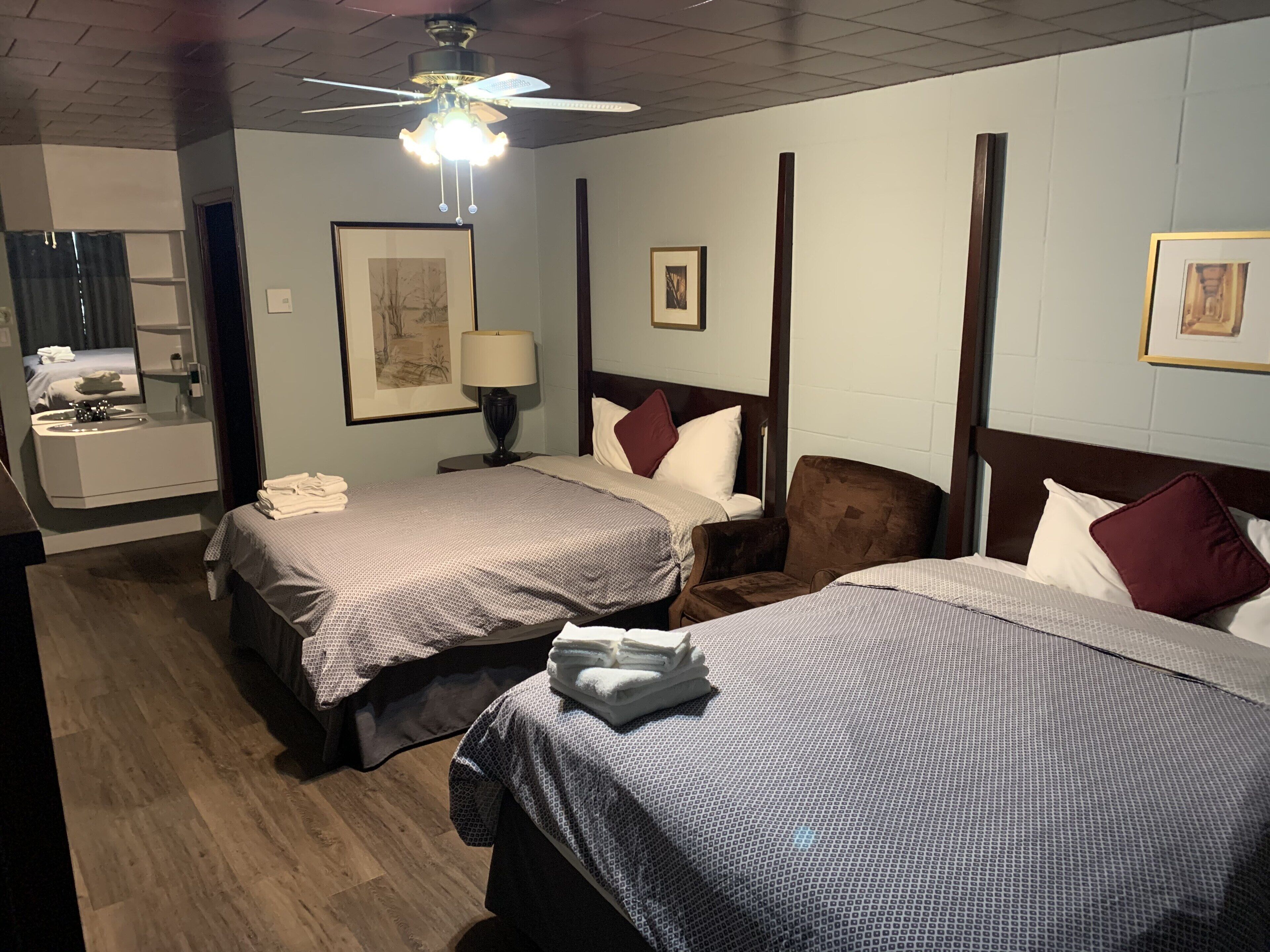A hotel room with two beds, a sitting area with a chair, a desk with a lamp, and a bathroom with a mirror and sink.
