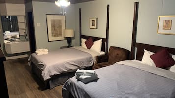 A hotel room with two beds, a sitting area with a chair, a desk with a lamp, and a bathroom with a mirror and sink.