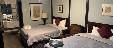 A hotel room with two beds, a sitting area with a chair, a desk with a lamp, and a bathroom with a mirror and sink.
