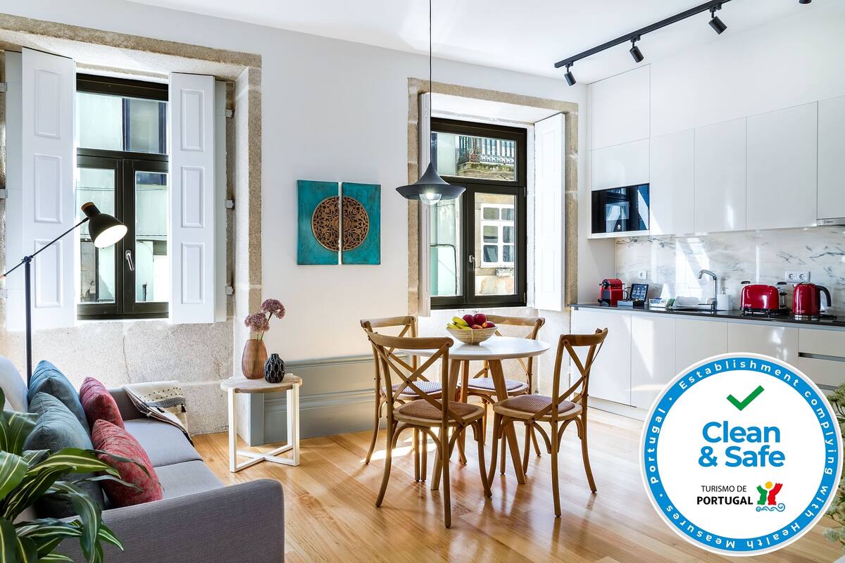 A modern living room with a sofa, dining table, and chairs, a kitchen with white cabinets, and a large window.