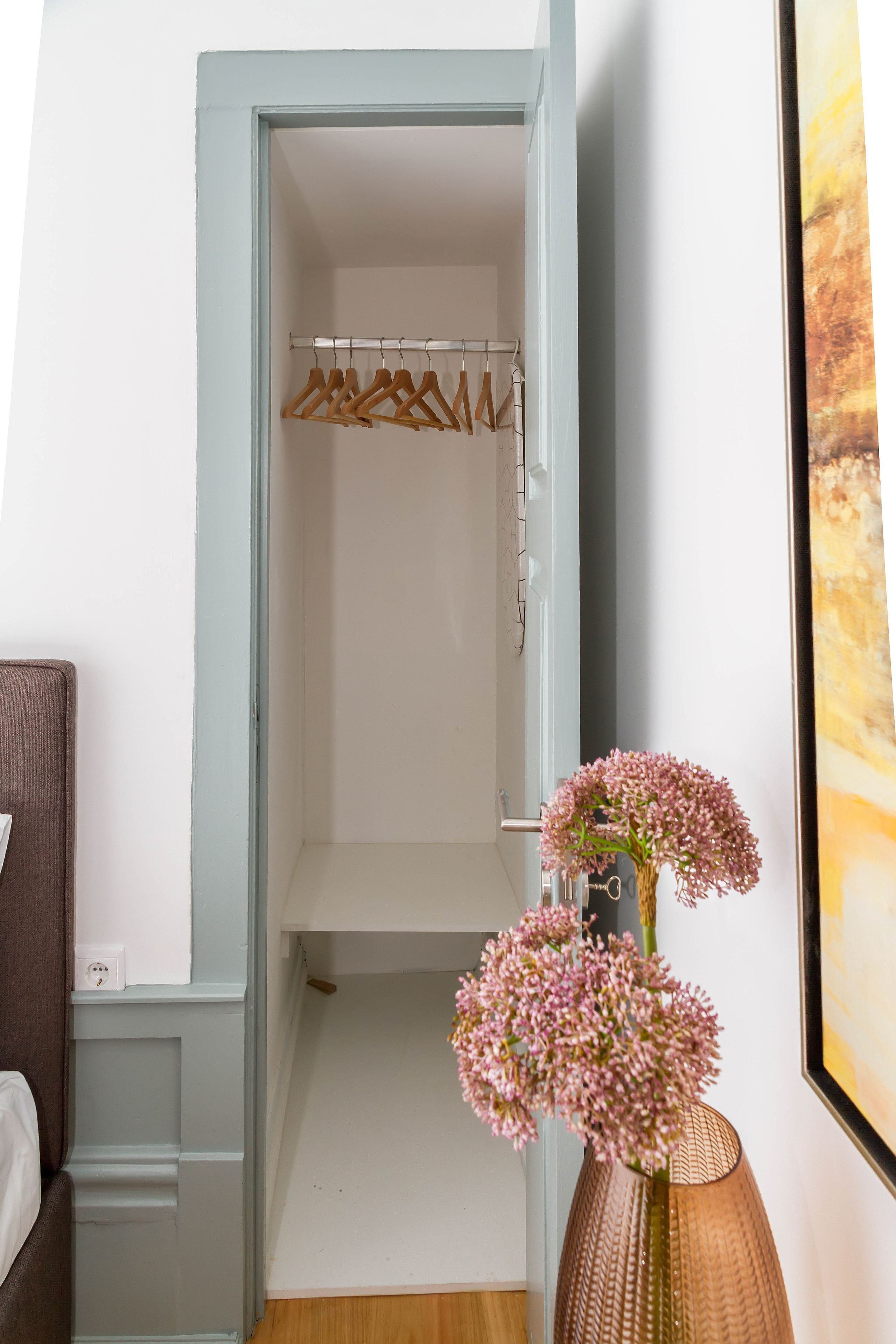 A hallway with a light blue door leading to a closet with wooden hangers. A vase with pink flowers is placed on the floor.