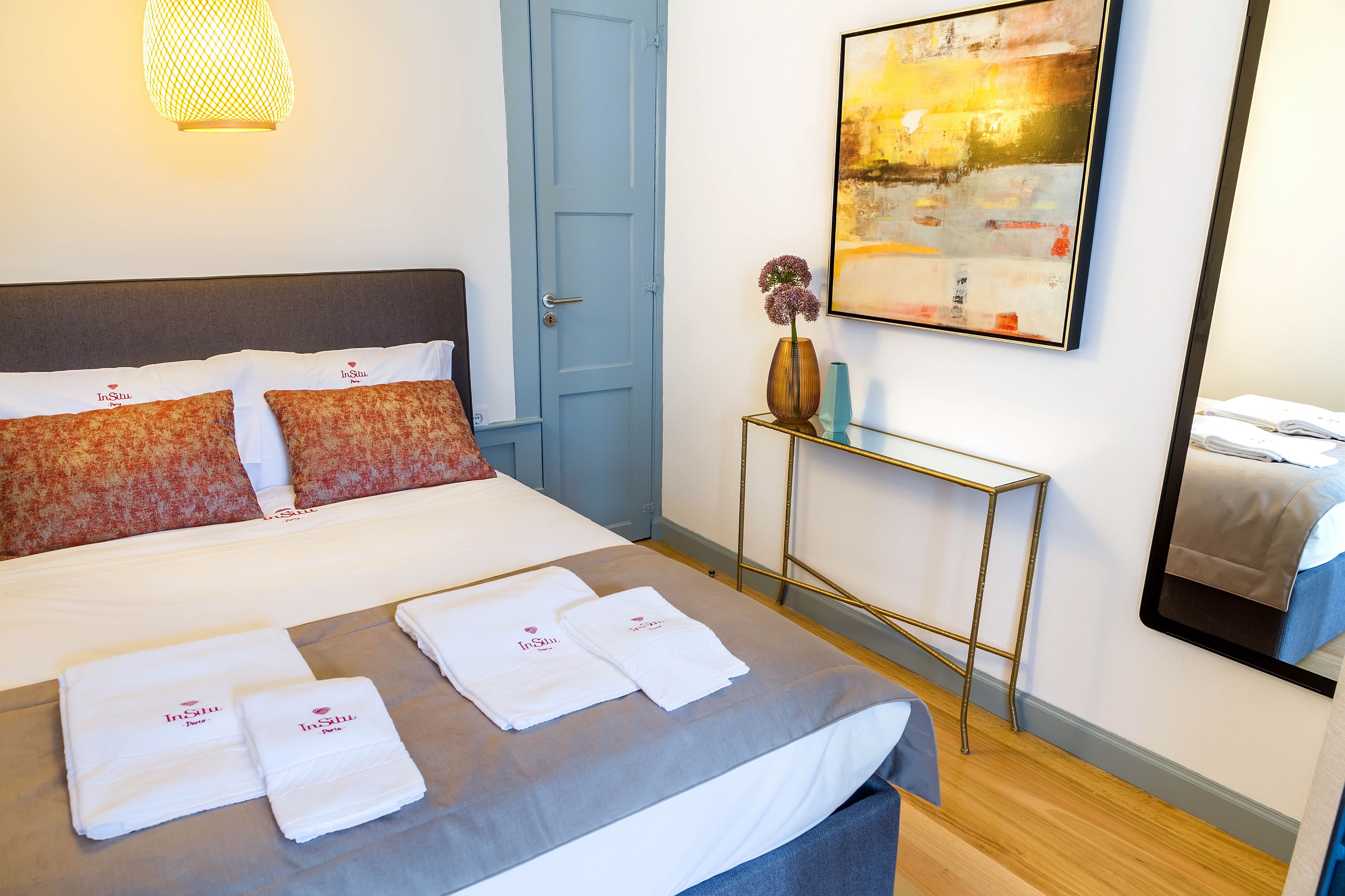 A hotel room with a bed, a bedside table with a vase and a lamp, a painting on the wall, and a mirror.
