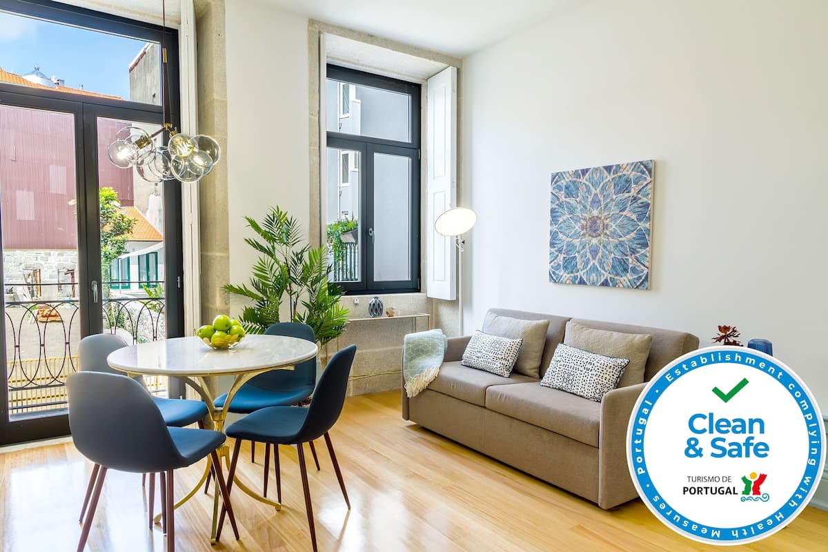 A modern living room with a sofa, a dining table with chairs, a potted plant, and a large window.